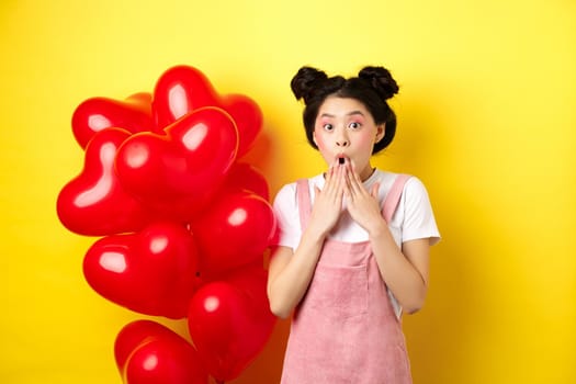 Valentines day concept. Impressed korean girl say wow, gasping and holding hands near opened mouth, staring at camera surprised, standing near romantic red heart balloons, yellow background.