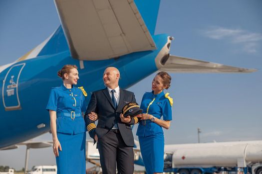 Excited pilot with two attractive stewardesses standing together in front of an airplane and smiling after landing. Aircraft, aircrew, occupation concept