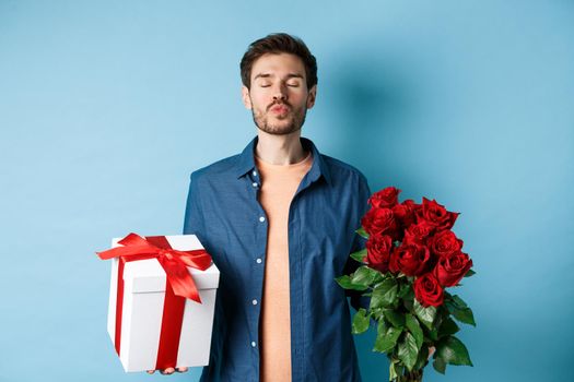 Love and Valentines day concept. Romantic man waiting for kiss, holding gift box and bouquet of red roses for lover on date, standing over blue background.