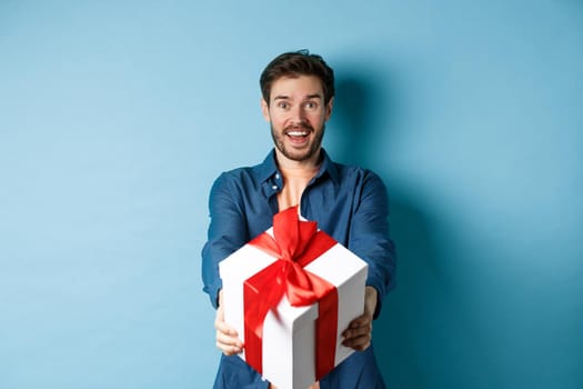 Valentines day. Excited handsome man giving gift box and wishing happy holiday. Romantic guy extending hands with present, standing againt blue background.