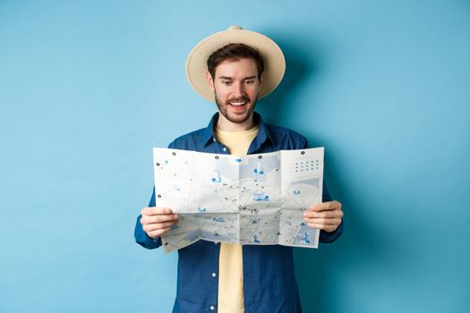 Cheerful tourist in summer hat planning travel route on vacation, looking at sighseeing map and smiling excited, standing on blue background.