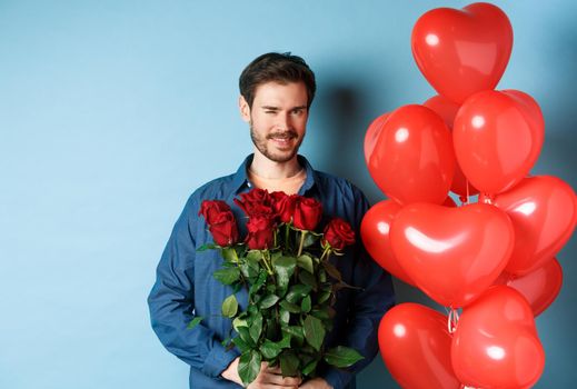 Romantic boyfriend winking and smiling, holding bouquet of flowers on Valentines day, standing near heart balloons for lover, blue background.