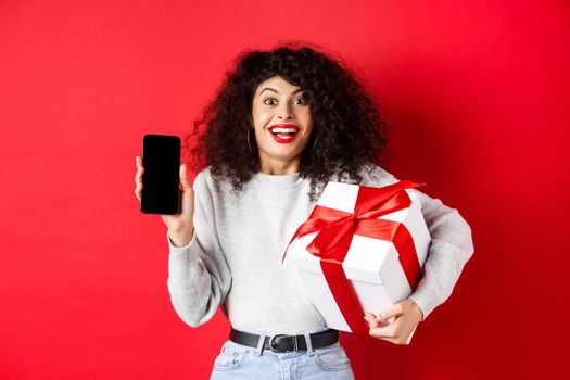 Valentines and lovers day. Excited smiling woman with curly dark hair, showing smartphone empty screen and holding surprise gift on holiday, showing online promo, red background.