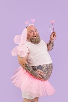 Happy bearded plus size male person in fairy costume with magic stick and wings poses on purple background in studio side view