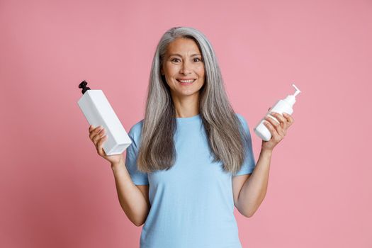 Pretty Asian woman with loose grey hair holds dispenser bottles of cosmetic products on pink background in studio. Mature beauty lifestyle