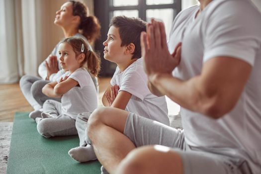 Calm woman is enjoying practice with husband and children and expressing namaste sign in lotus position