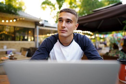 Young man sitting at table and typing on laptop keyboard while working in outdoor cafe, close up