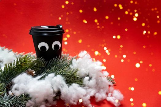 Black coffee cup with plastic cap and googly eyes among green braches with knar and snow on red background with lights, bokeh style, new year concept, place for the text.