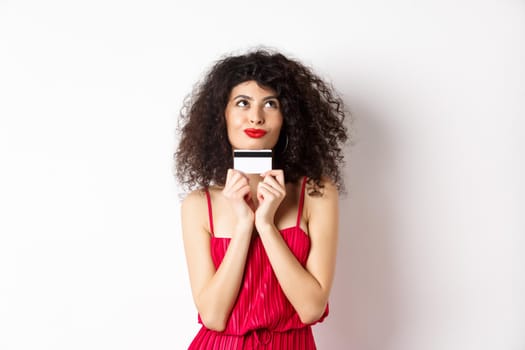 Silly young woman looking romantic, holding plastic credit card and thinking of shopping, standing over white background. Copy space