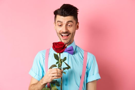 Valentines day and romance concept. Happy boyfriend looking at red rose while waiting for lover in bow-tie, standing on pink romantic background.