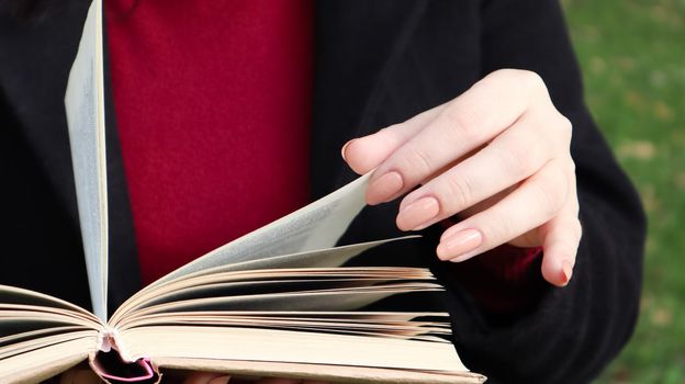 Girl reading a book in the park. Female hands flipping pages of paper book outdoors. The student is preparing for the exam. Literary leisure in nature. Close-up, copy space