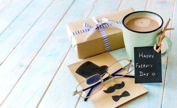 Happy father's day message written by child's hands on tag with handmade greeting card, glasses, giftbox and cup of coffee on blue wooden background