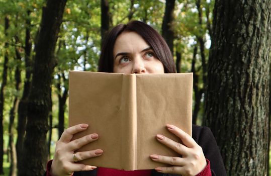 Portrait of a young woman with an open untitled book held close to her face. Brunette covering half of her face with a book in the park on a sunny day. Education and people concept, selective focus