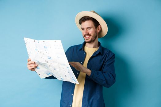 Handsome smiling tourist in straw hat looking at map, choosing travel road, planning a vacation, standing on blue background.