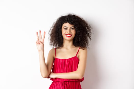 Beautiful lady with curly hair, wearing elegant red dress and showing number three, making order, standing over white background.