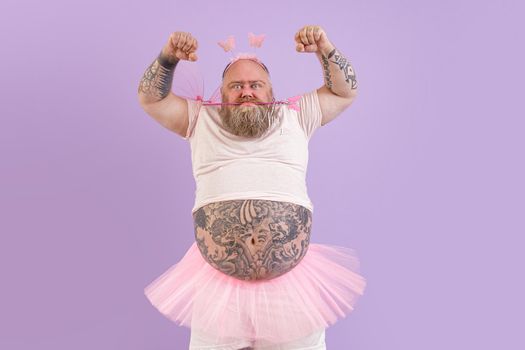 Mature plus size man with tattoos wearing fairy costume shows muscles holding magic stick in teeth on purple background in studio