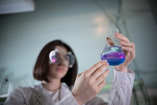 Chemical laboratory. Young woman holding a flask with blue and purple exfoliating liquid in it. Flask in focus