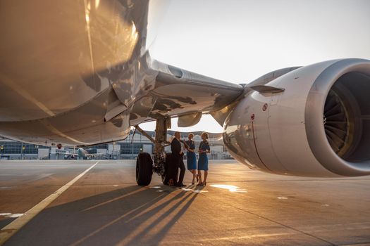 Airplane near the terminal in an airport at the sunset. Pilot and two stewardesses standing together and talking after landing or before departure. Aircraft, aircrew, occupation concept