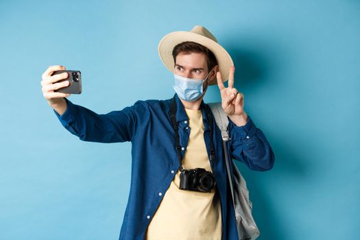 Covid-19, pandemic and travel concept. Happy and positive guy in tourist hat taking selfie and showing peace sign, posing near sightseeing, blue background.