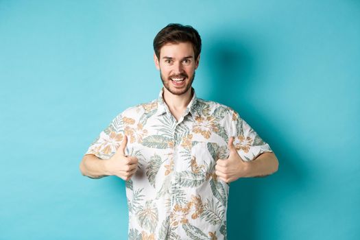 Handsome tourist showing thumbs up and say yes, praising good tourism agency, wearing hawaiian shirt, standing on blue background.
