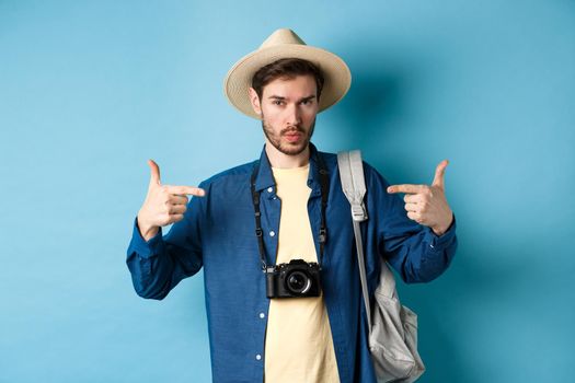 Confident and ready young man pointing at himself, self-promoting or show-off, going on summer vacation, wearing straw hat, holding backpack with camera, blue background.