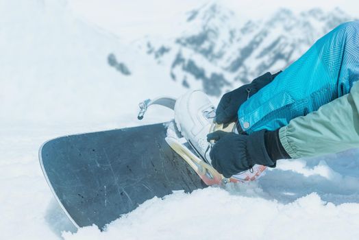 Unrecognizable man putting on his snowboard and tightening the straps on background of snowy mountains in winter, side view