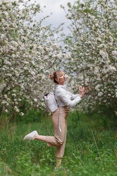 Beautiful young blonde woman in white shirt with backpack posing under apple tree in blossom and green grass in Spring garden