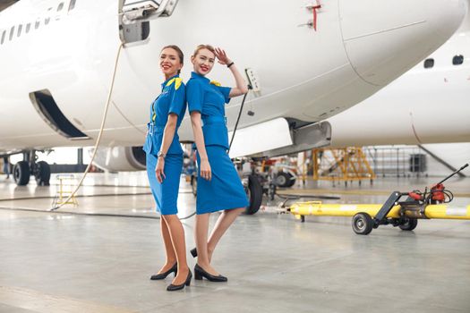 Full length shot of two pretty air stewardesses in bright blue uniform smiling at camera, posing together in front of passenger aircraft in hangar at the airport. Occupation concept