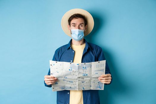 Covid-19, pandemic and travel concept. Tourist in medical mask look surprised, holding map, standing on blue background.