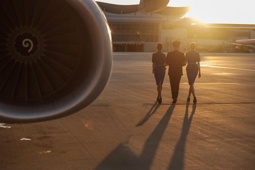 Full length shot of professional pilot walking together with two female flight attendants in blue uniform in terminal at sunset. Aircraft, aircrew concept. Back view