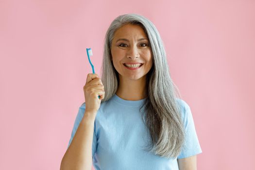 Cheerful middle aged Asian woman with grey hair in blue t-shirt holds toothbrush standing on pink background in studio. Oral cavity hygiene