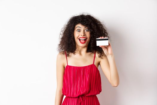 Excited smiling woman in red dress, showing plastic credit card and looking happy, standing over white background. Copy space