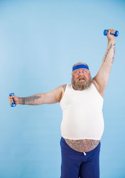 Shocked mature obese man with beard does exercises with dumbbells training hard on light blue background in studio