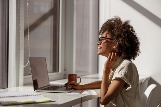 Side view of happy pretty woman in glasses typing on laptop during sunny day in coworking