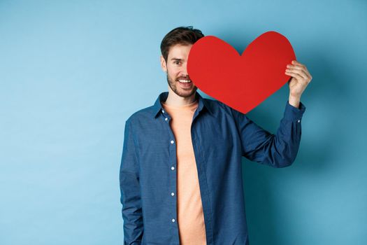 Happy man showing valentines heart and smiling, make romantic gift on lovers day, standing over blue background.
