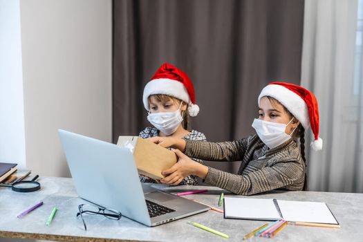 Christmas greetings online. two little girls in medical masks a laptop. Shows gifts, online purchases to the camera. Greeting video calls to friends, relatives and parents