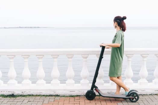 Modern girl in dress standing with an electric scooter on embankment and looking at the sea in summer.