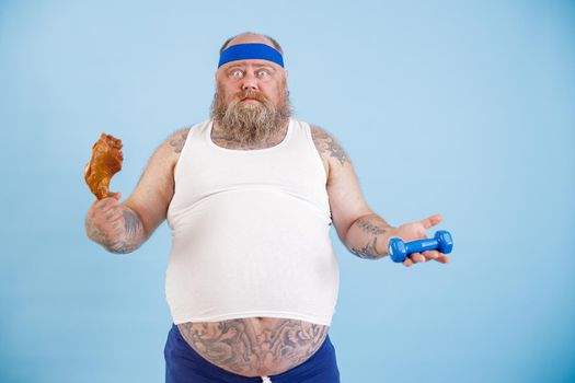 Confused mature man with overweight in tracksuit chooses between chicken leg and dumbbell standing on light blue background in studio