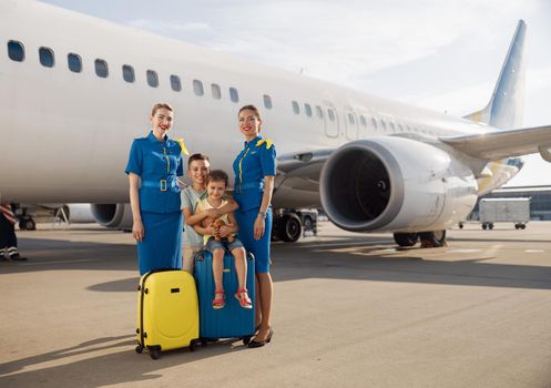 Full length shot of happy two kids sitting on their luggage in front of big airplane on a daytime and smiling at camera together with beautiful stewardesses. Family, traveling, vacation concept