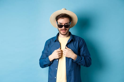 Handsome young man going on vacation, adjusting shirt, wearing sunglasses and straw hat. Concept of summer holiday.