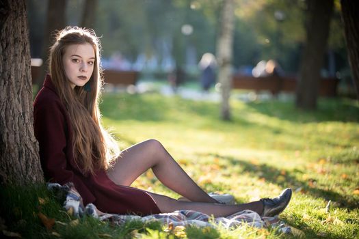 Beautiful girl with dark lipstick sitting in the park near the tree. Natural daylight.