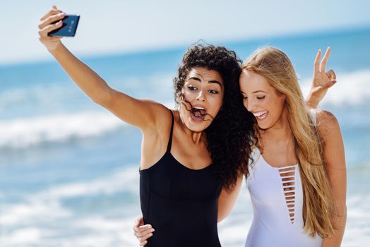 Two young women taking selfie photograph with smart phone in swimsuits on a tropical beach.