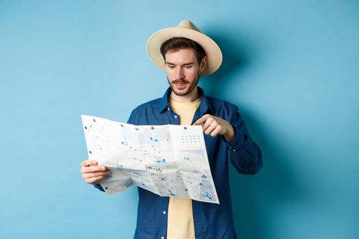 Handsome guy choosing place to go, pointing at sighseeing map on vacation, travelling on summer holidays, wearing straw hat and shirt, blue background.