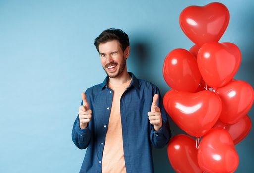 Happy valentines day. Cheeky boyfriend winking and pointing fingers at camera, standing with red romantic balloons on blue background. Copy space