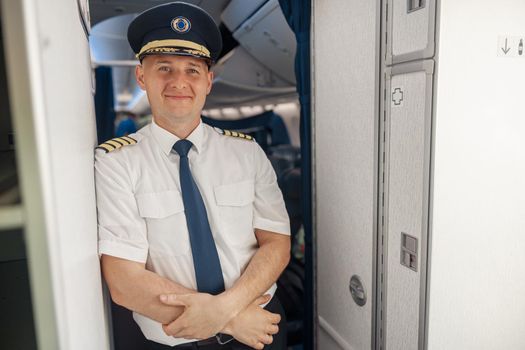 Happy male pilot in uniform and hat keeping arms crossed and smiling at camera while standing inside of the airplane. Transportation, aircrew concept