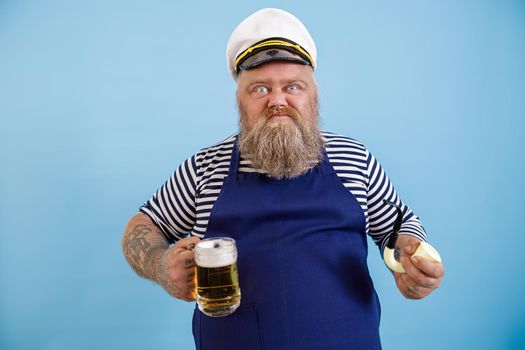 Funny aggressive obese man in sailor costume with apron holds smoking pipe and glass mug of delicious beer on light blue background in studio