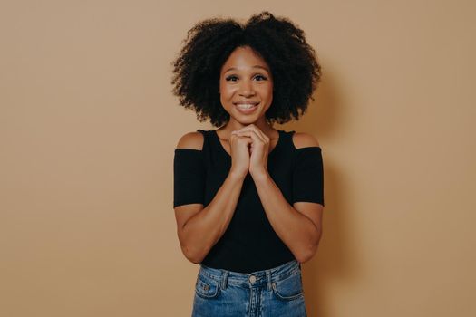 Mixed race young woman holding hands in prayer gesture, smiling with eyes opened isolated over dark beige background with copy space. African lady posing in studio, hope and positive emotions concept