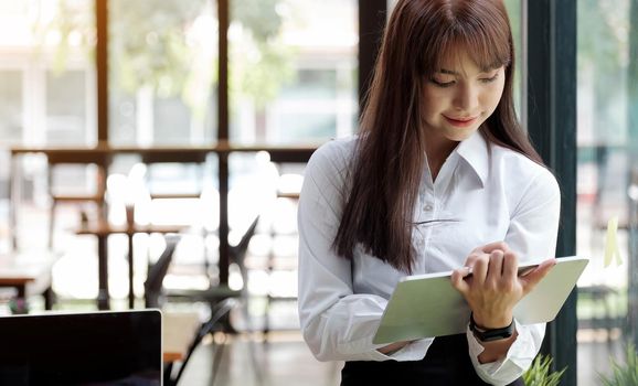 Young adult business lady in white holding digital tablet in hands and looking at digital tablet . Businesswoman holding device standing on office.