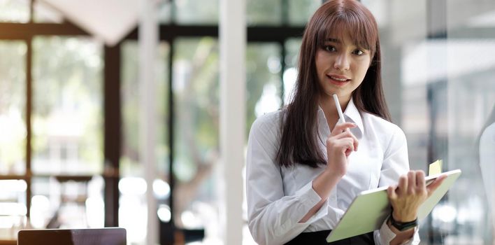 An elegant businesswoman using a tablet in the back of her office is standing looking at the camera.