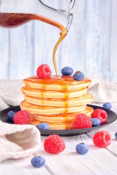 Delicious pancakes with fresh blueberries, raspberries are poured with sweet maple syrup on a light wooden background.